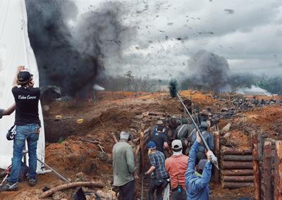 color image of battle scene on film set; black cloud of smoking rising on L, behind white screen and crew member facing away from viewer; reinforced trench dug into field at center and R, with more explosions in battlefield in background; production crew with equipment in trench foreground, with people dressed as soldiers further back in trench, firing muskets and facing battlefield