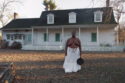 color image of a topless Black woman wearing a long white skirt with two pairs of baby shoes tied around her waist and holding a cast iron skillet in her PL hand; white Colonial-style house with green shutters behind woman