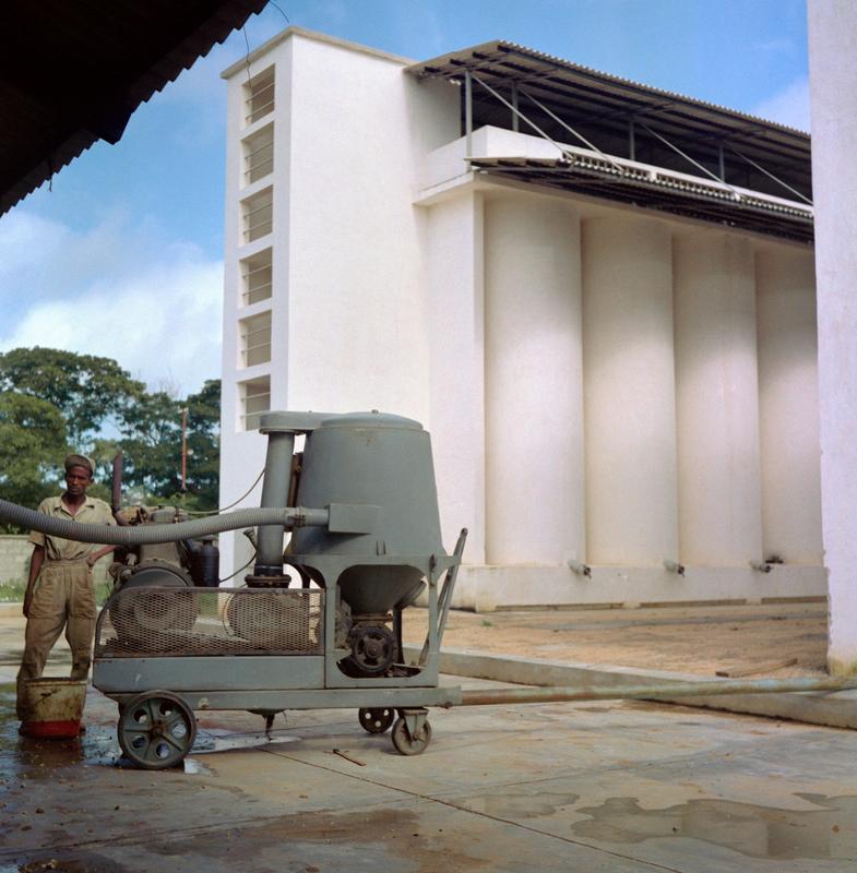 Color photograph of a figure operating a green machine in front of a white grain storage building